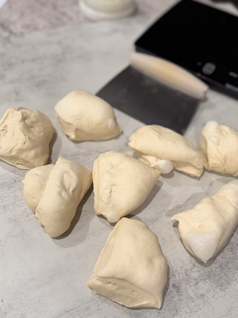 Several pieces of raw dough rest on a floured countertop, with a metal dough scraper and a digital scale visible in the background.