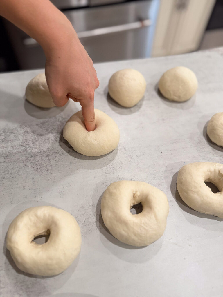 A hand presses a finger into a round ball of dough on a countertop, with other dough balls and ring-shaped dough pieces nearby.