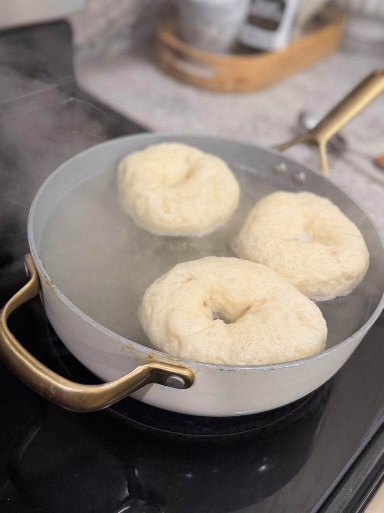 Three bagels are boiling in a shallow pan of water on a stovetop, with steam rising and kitchen items visible in the background.