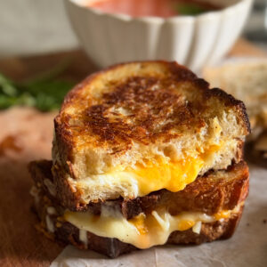 A close-up of two stacked grilled cheese sandwiches on parchment paper with a bowl of tomato soup in the background.