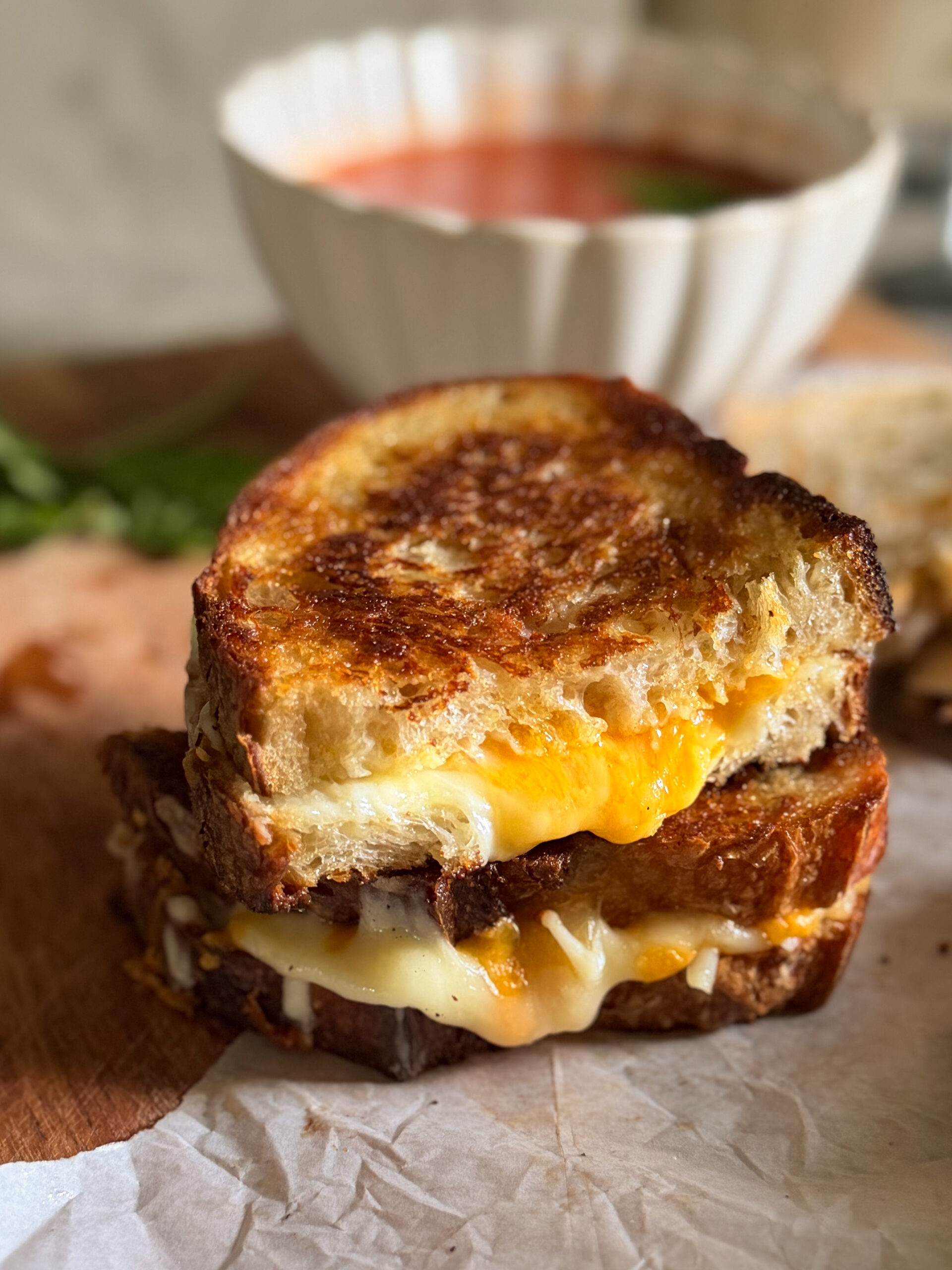 A close-up of two stacked grilled cheese sandwiches on parchment paper with a bowl of tomato soup in the background.