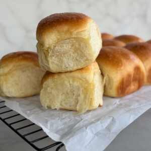Three golden brown Hawaiian sourdough rolls are stacked on top of each other on a cooling rack lined with parchment paper, with more rolls in the background.