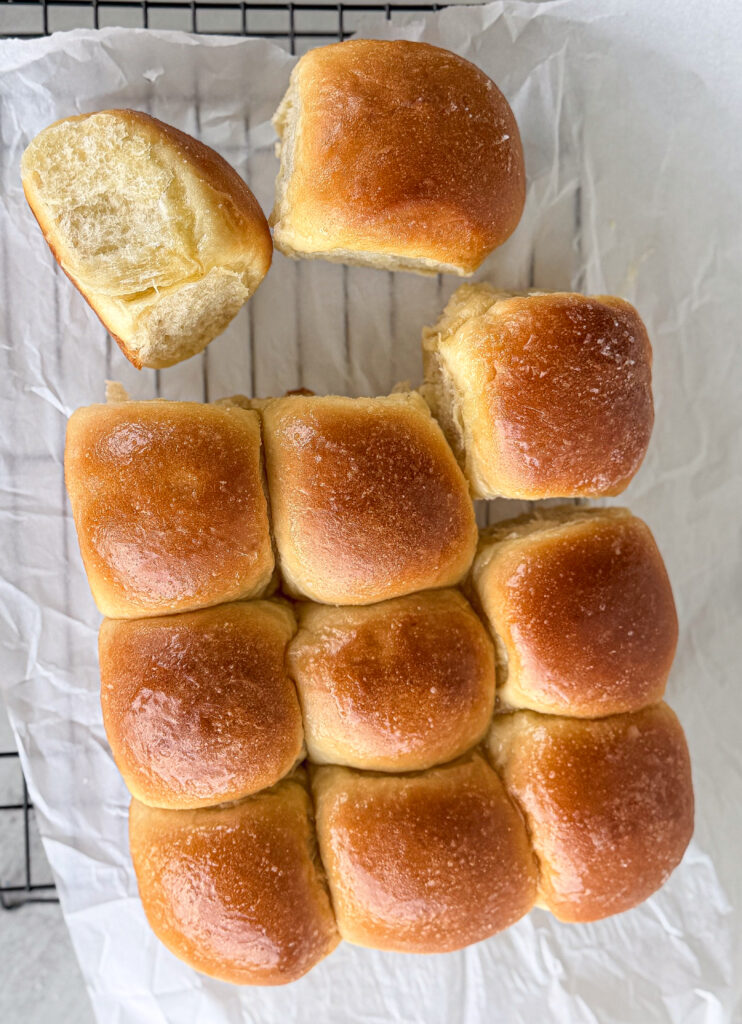 Nine golden brown bread rolls on parchment paper, with one roll partially separated and another pulled apart to show its soft interior.