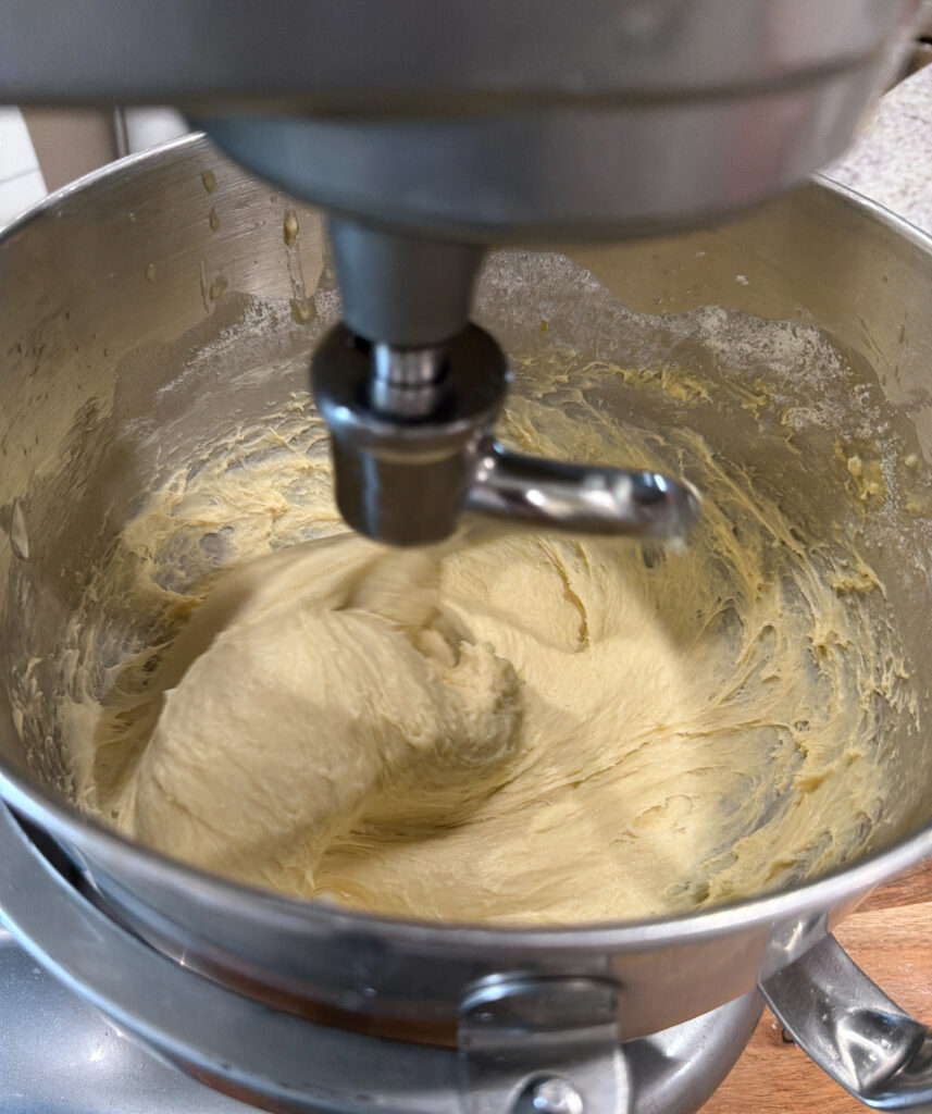 A stand mixer with a dough hook attachment is mixing a thick, yellow dough in a stainless steel bowl.