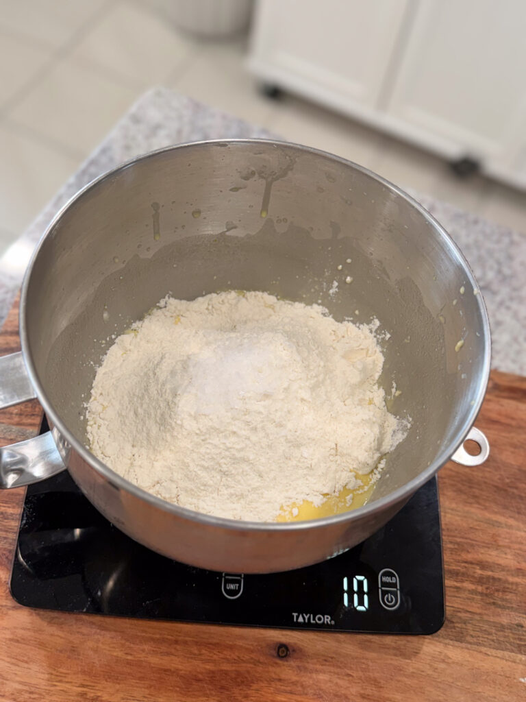 A metal mixing bowl with flour and other ingredients sits on a digital kitchen scale displaying a weight of 10 grams, on a wooden surface.
