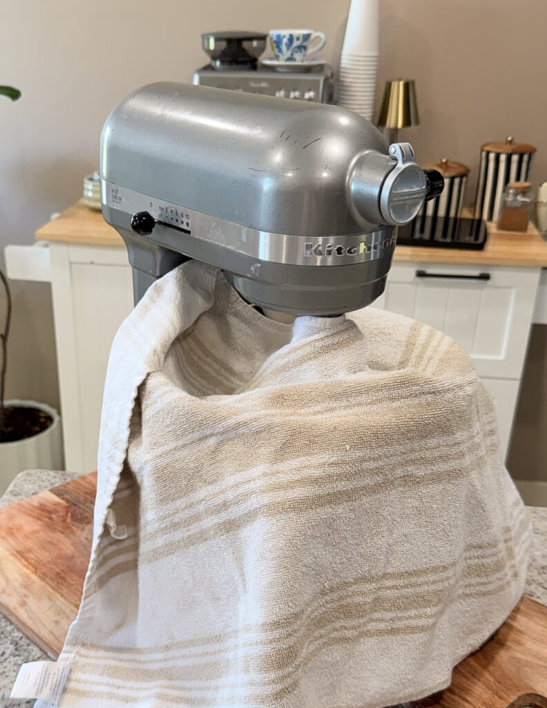 A stand mixer covered with a beige striped towel sits on a wooden board in a kitchen.