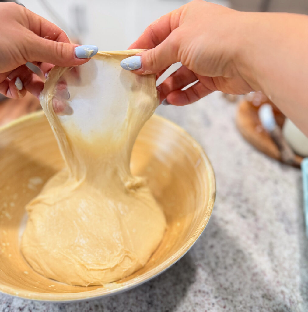 Hands stretch dough in a bowl to check gluten development; bowl is on a countertop with baking items partially visible in the background.