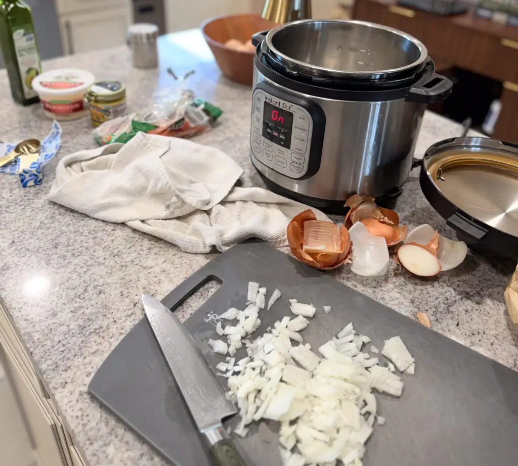 Chopped onions and a knife on a cutting board sit next to an Instant Pot and onion peels on a kitchen counter with various ingredients and utensils around.