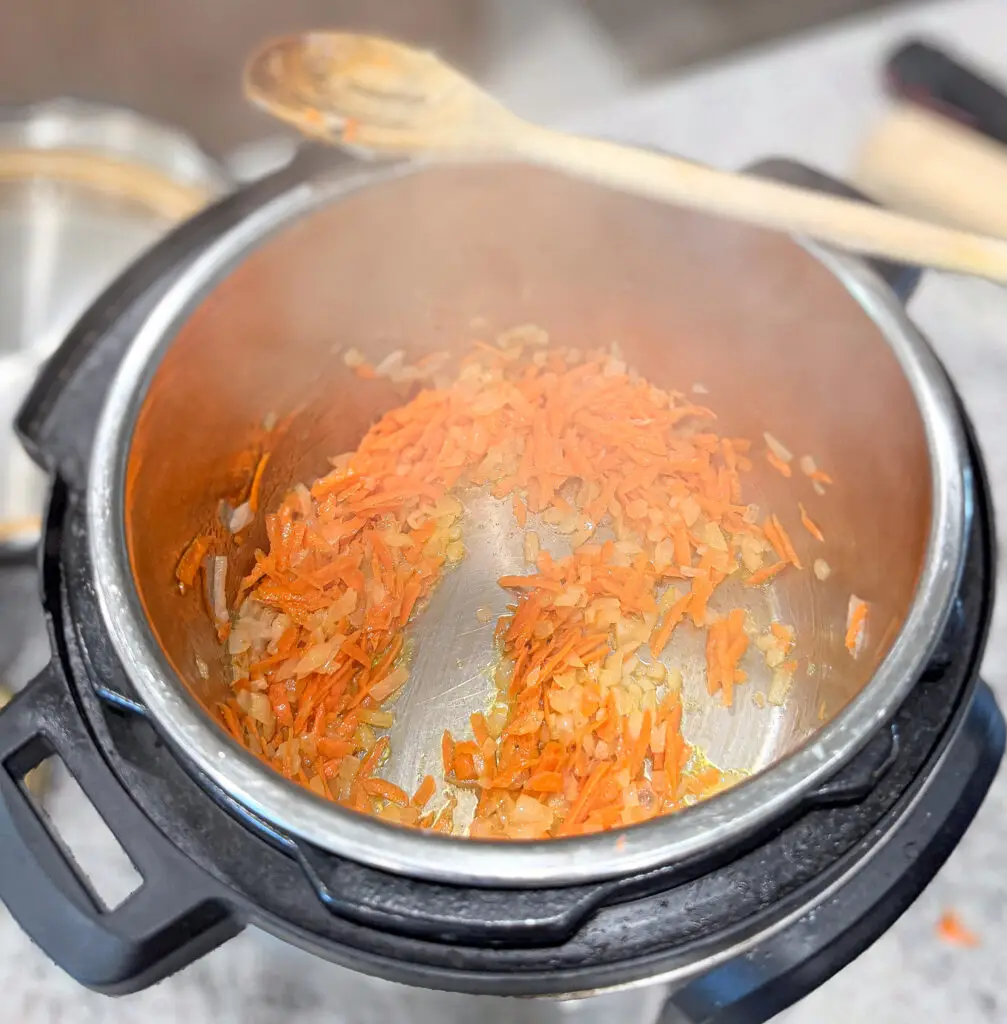 Chopped onions and grated carrots sautéing in an Instant Pot with a wooden spoon resting on top.