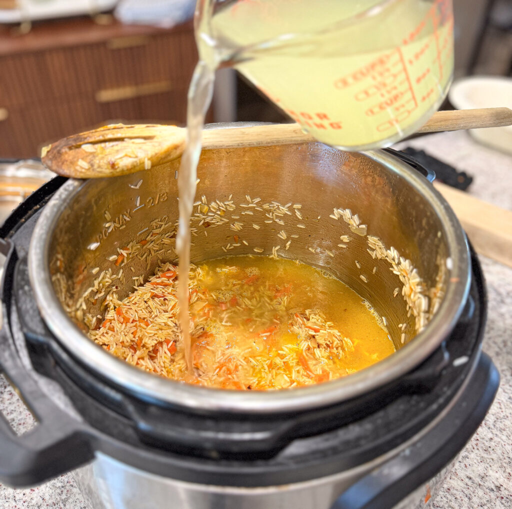 A measuring cup pours liquid into an Instant Pot filled with rice, vegetables, and seasonings on a kitchen counter.