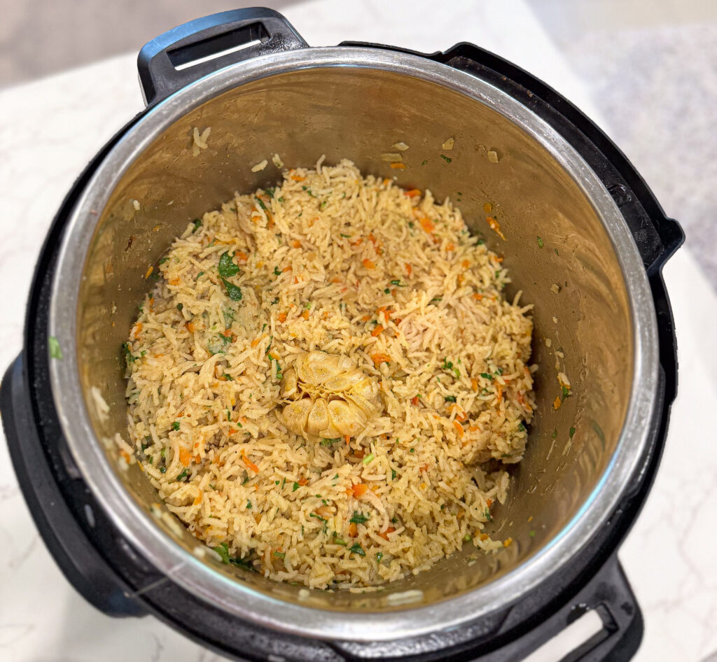 Cooked vegetable rice with a whole head of garlic in the center, shown inside a stainless steel inner pot of an electric pressure cooker.