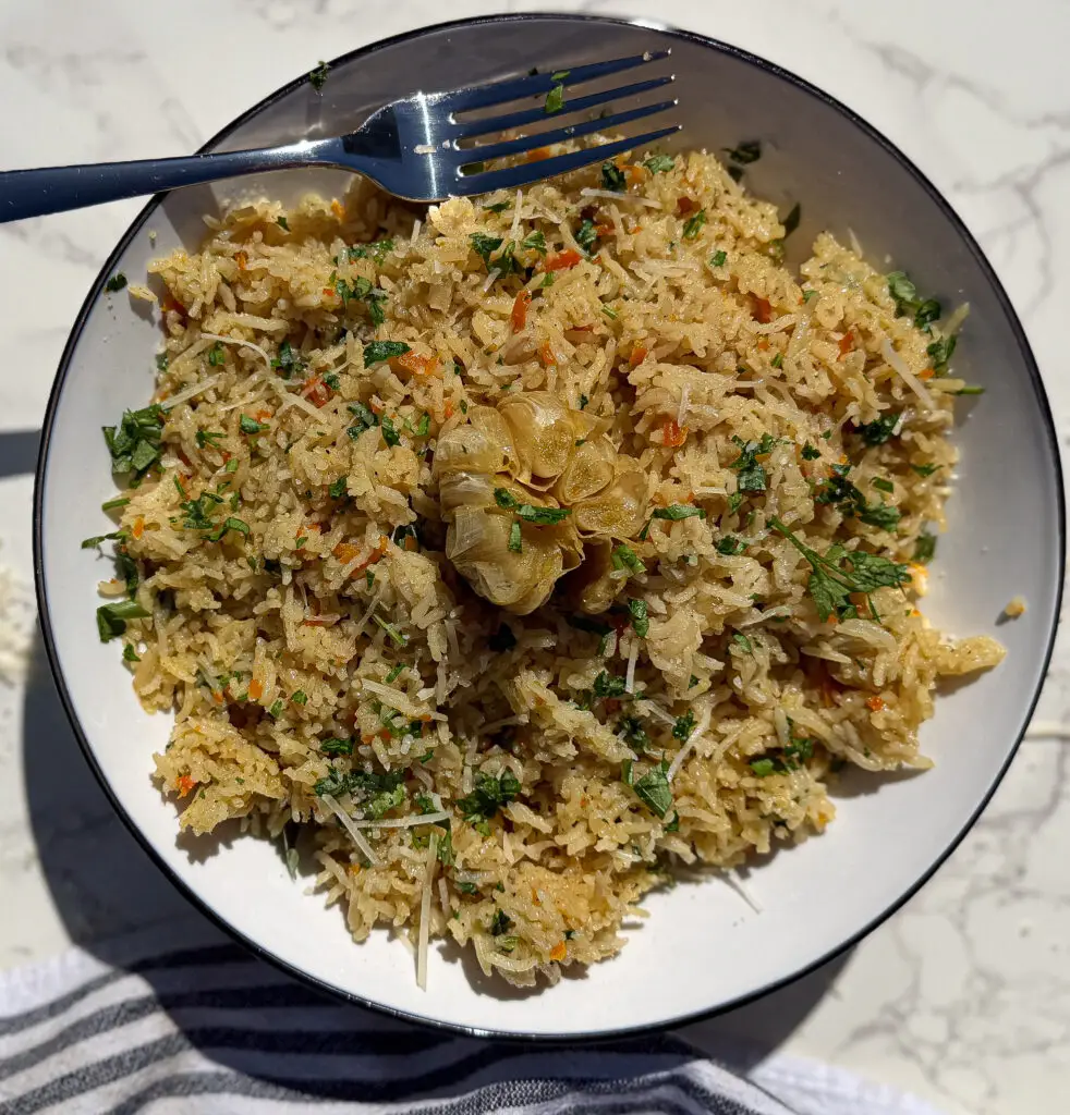 A bowl of vegetable fried rice garnished with fresh herbs and a whole roasted garlic bulb, with a fork resting on the edge, on a marble surface.