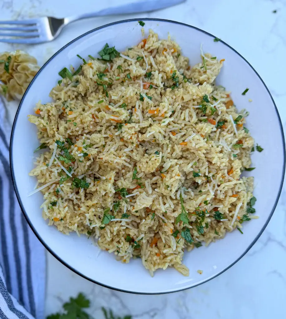 A white bowl filled with cooked vegetable rice, garnished with chopped herbs and shredded cheese, placed on a marble surface with a fork and striped napkin nearby.
