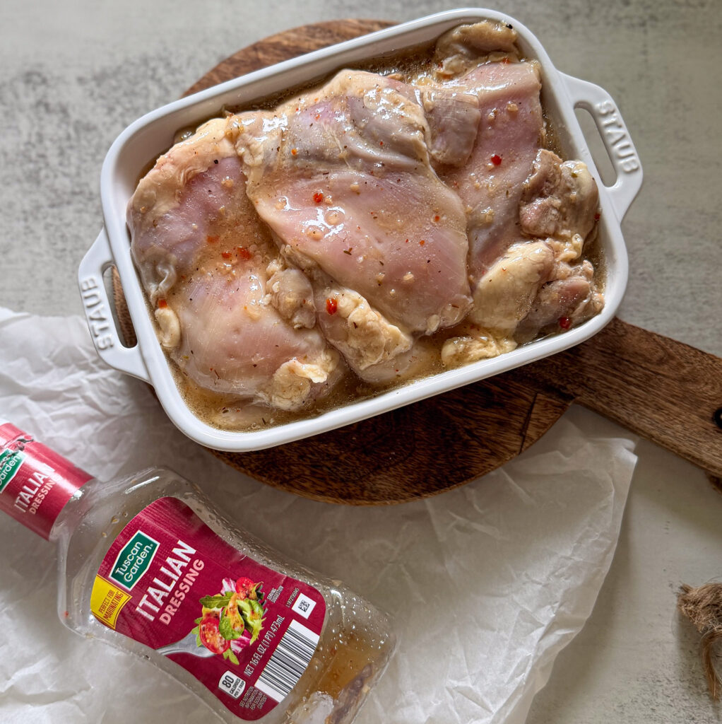 Raw chicken marinating in Italian dressing inside a white ceramic dish, with an empty bottle of Italian dressing and a wooden cutting board nearby.