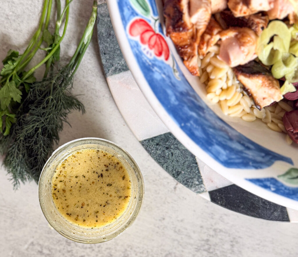 A glass of herb dressing sits next to a bowl of pasta salad with salmon, olives, and celery, with fresh herbs placed nearby on a counter.
