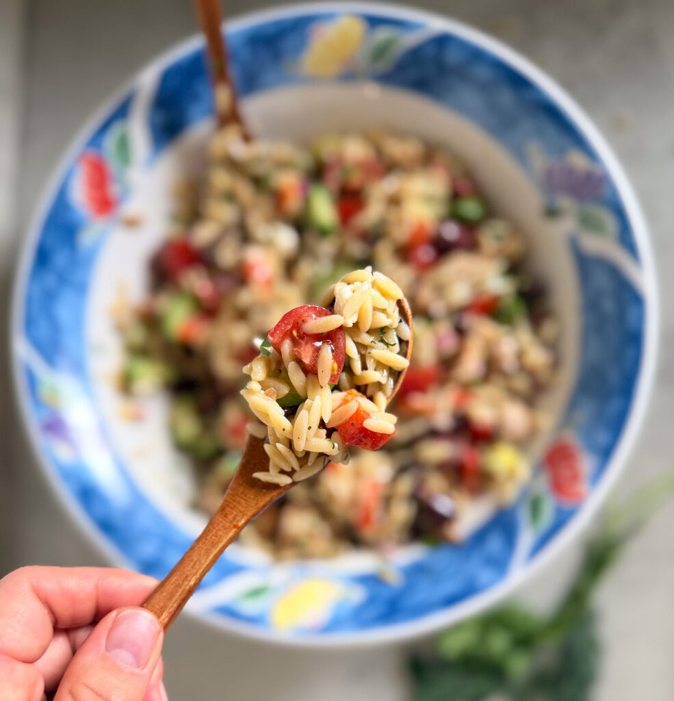 A hand holds a wooden spoon with a portion of orzo salad containing vegetables over a large bowl filled with the same salad.
