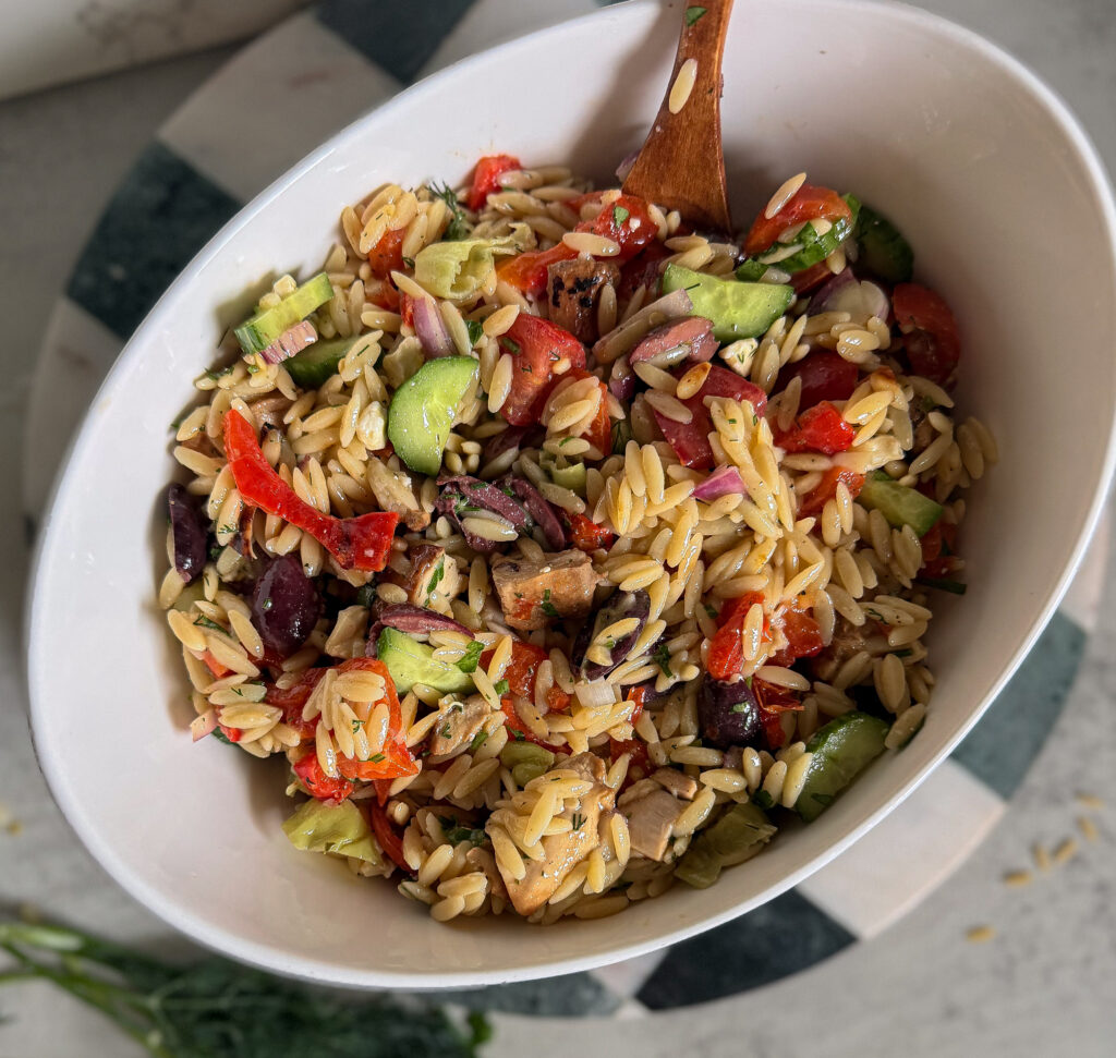 Oval bowl filled with orzo pasta salad mixed with chopped vegetables, olives, and herbs, with a wooden spoon resting inside.