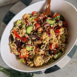 Oval bowl filled with orzo pasta salad mixed with chopped vegetables, olives, and herbs, with a wooden spoon resting inside.