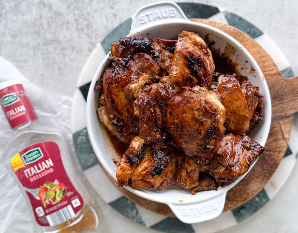 A white dish filled with grilled, glazed chicken pieces sits on a wooden board, with a bottle of Italian salad dressing nearby.