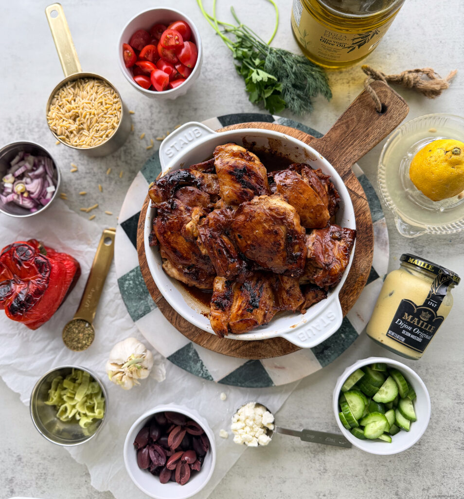 Grilled chicken pieces in a white bowl surrounded by fresh vegetables, olives, mustard, orzo, and various seasonings on a kitchen counter.