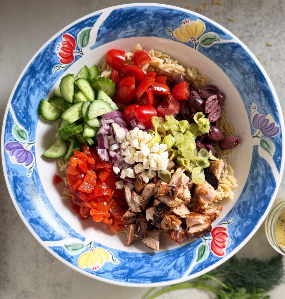 A bowl of orzo pasta topped with grilled chicken, feta cheese, chopped tomatoes, cucumbers, red onion, roasted red peppers, kalamata olives, and pepperoncini, arranged in sections.