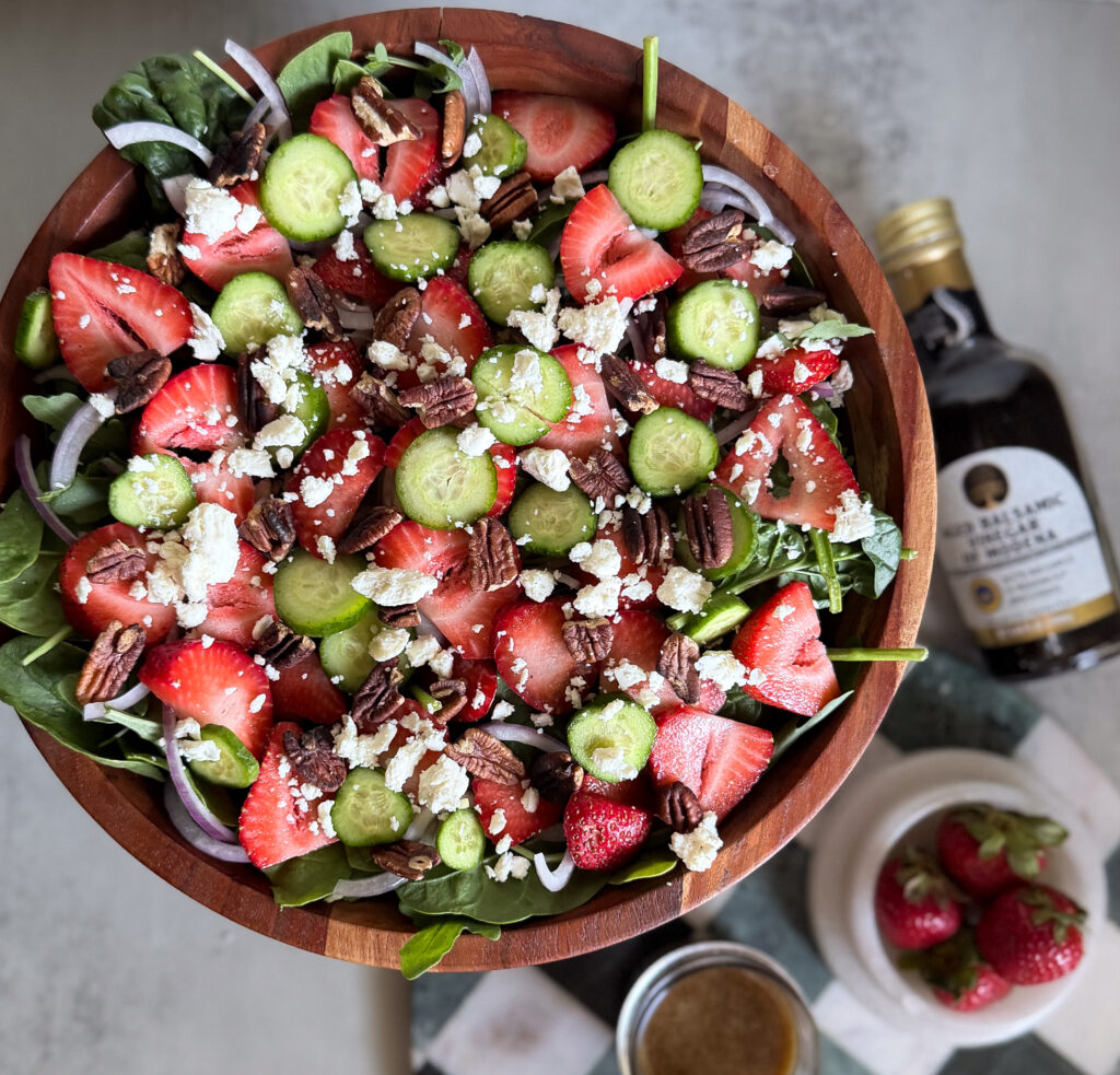 A wooden bowl filled with spinach salad topped with strawberries, cucumber slices, pecans, red onions, and crumbled cheese. A bottle of balsamic vinegar sits beside the bowl.