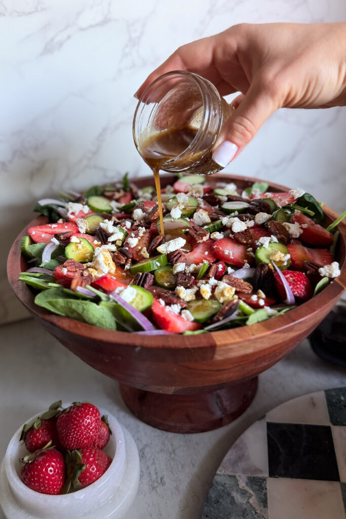 A hand pours dressing from a small jar onto a salad with spinach, strawberries, grapes, feta cheese, and nuts in a wooden bowl. A small bowl of strawberries is nearby.