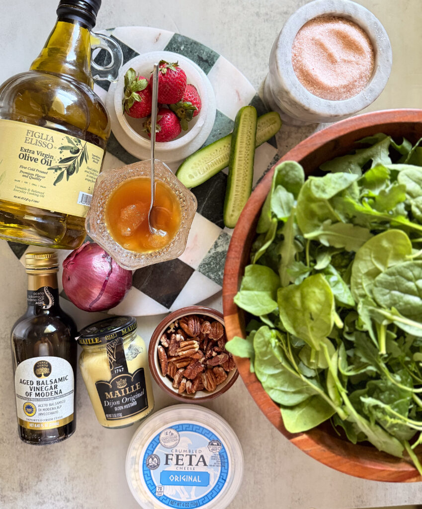 A bowl of fresh spinach sits beside containers of feta cheese, pecans, Dijon mustard, olive oil, balsamic vinegar, honey, salt, strawberries, cucumber, and a red onion.