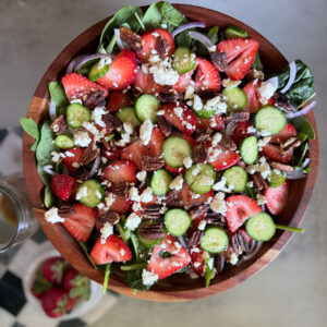 A wooden bowl filled with salad made of spinach, sliced strawberries, cucumber, red onion, pecans, and crumbled cheese.