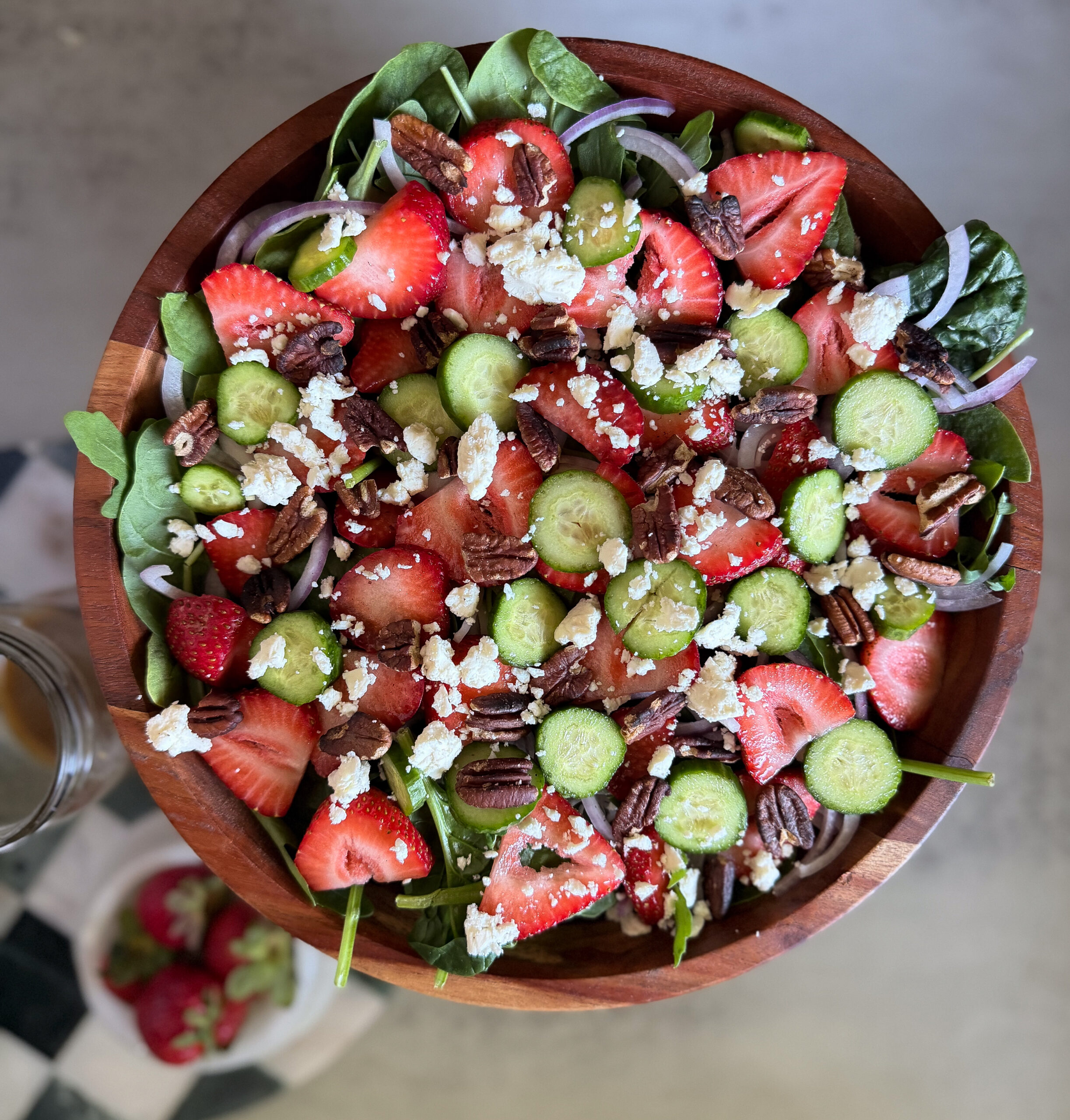 A wooden bowl filled with salad made of spinach, sliced strawberries, cucumber, red onion, pecans, and crumbled cheese.