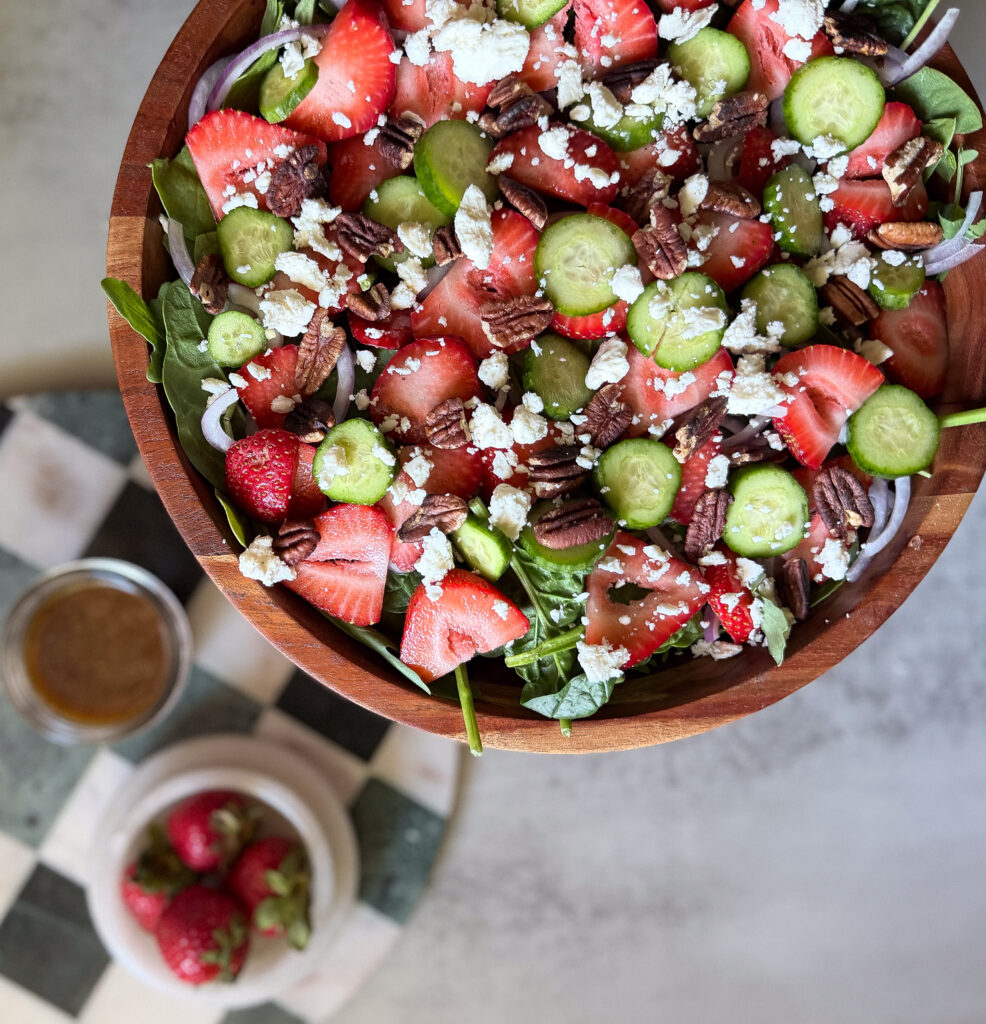 A wooden bowl filled with salad containing spinach, sliced strawberries, cucumbers, pecans, and crumbled cheese, with a jar of dressing and a small bowl of strawberries nearby.