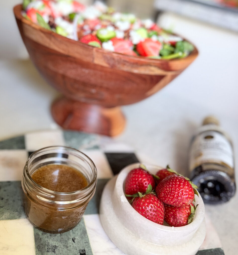 A wooden bowl of salad with strawberries and cheese, a jar of dressing, a bowl of strawberries, and a bottle of balsamic vinegar on a countertop.