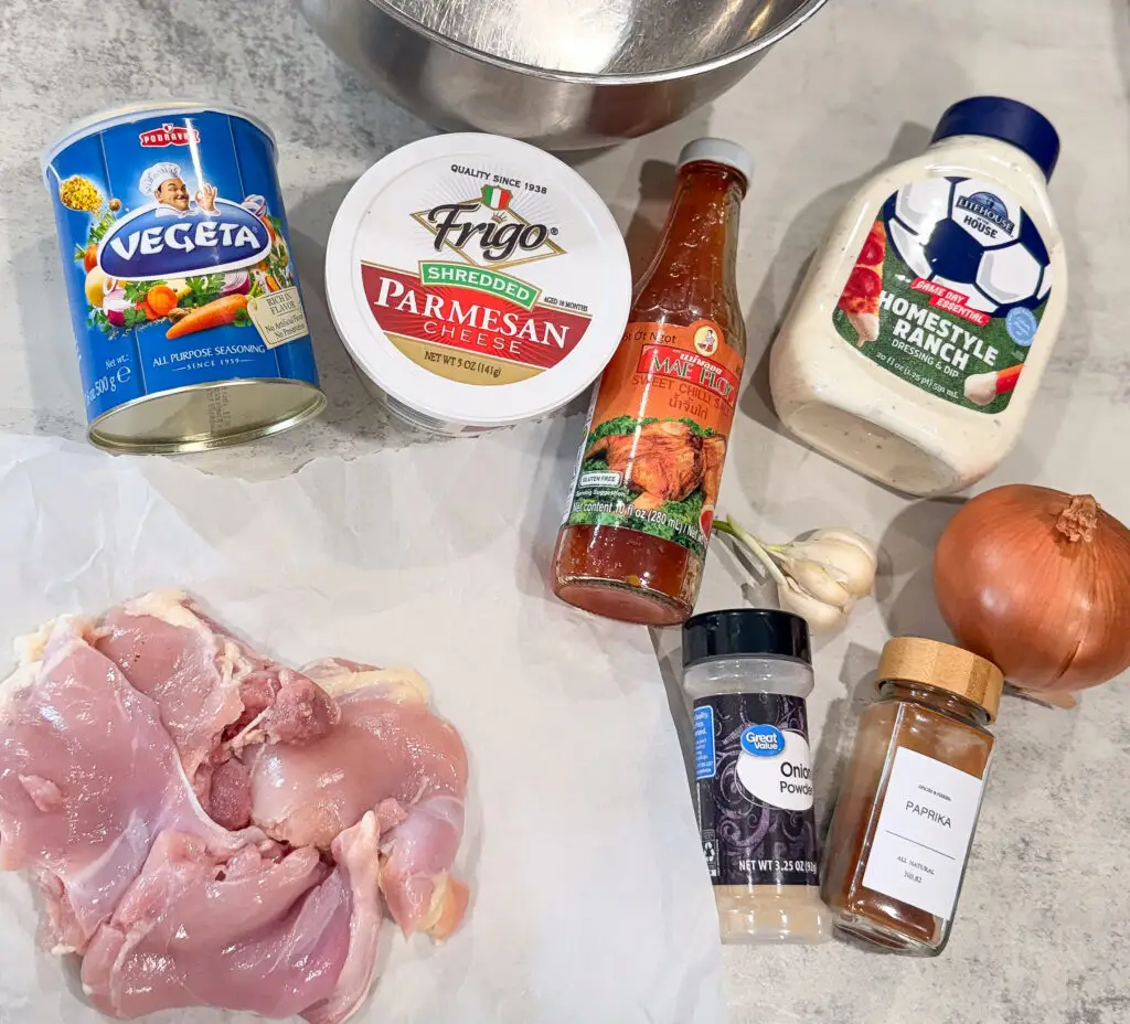 Raw chicken thighs, assorted seasonings, shredded parmesan cheese, buffalo sauce, ranch dressing, garlic cloves, and a whole onion arranged on a countertop.