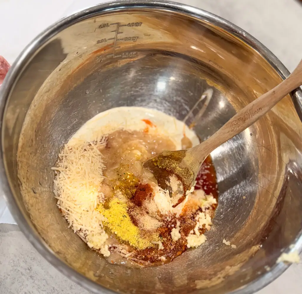 A metal mixing bowl contains grated cheese, spices, breadcrumbs, sauce, and other ingredients being stirred with a wooden spoon.