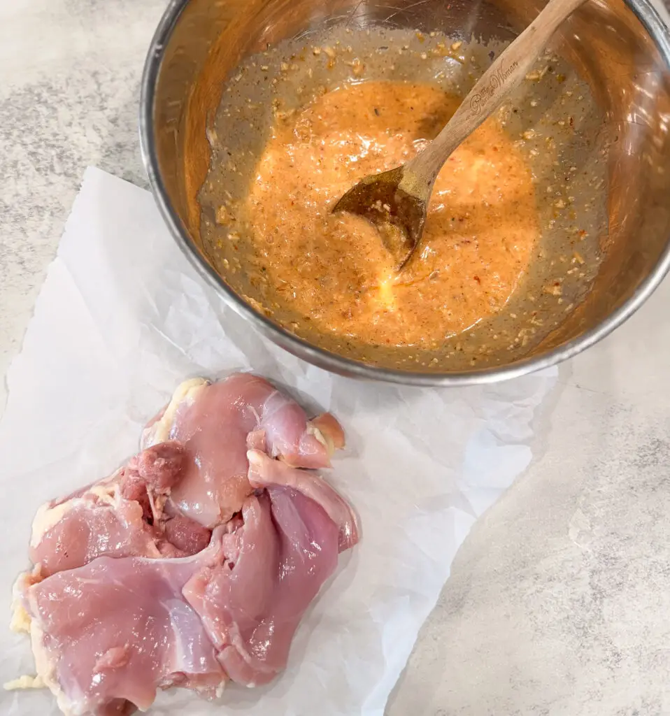 Raw chicken thighs on parchment paper next to a metal bowl containing a seasoned marinade with a wooden spoon.