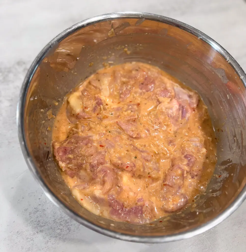 Raw pieces of meat marinating in a creamy, seasoned mixture inside a stainless steel bowl on a light-colored surface.
