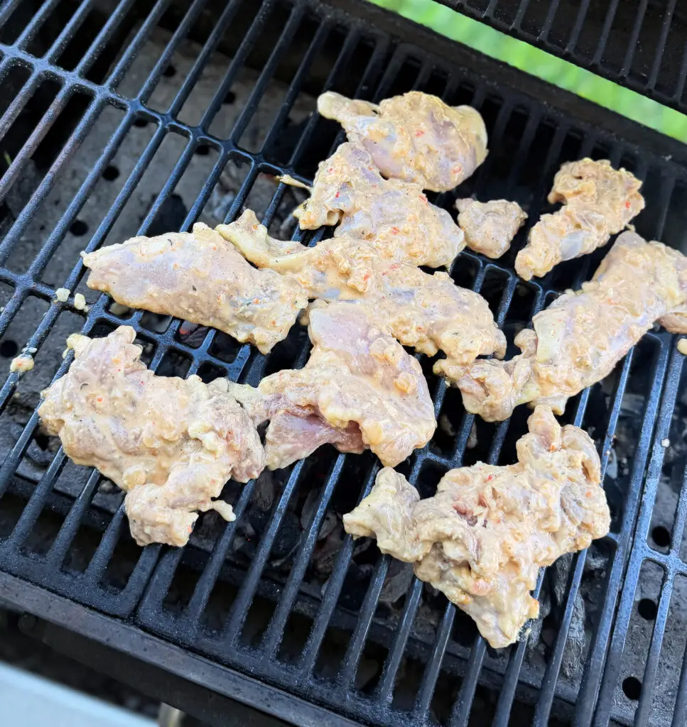 Marinated pieces of raw meat are spread out on a metal grill over open flames, ready to be cooked outdoors.