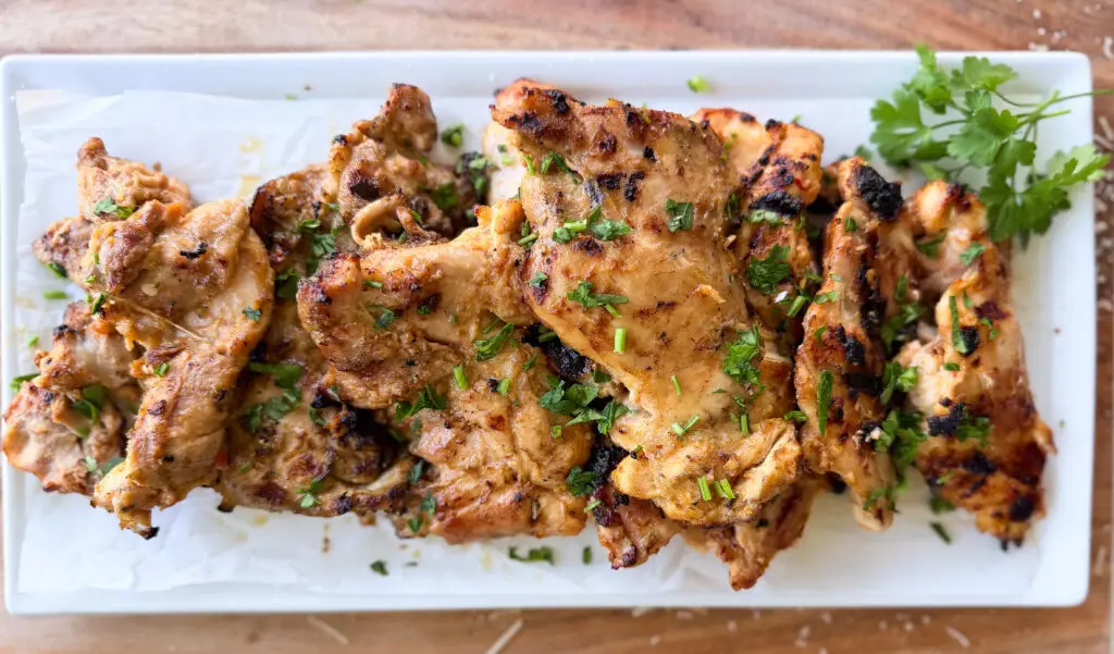 Grilled marinated chicken thighs garnished with chopped herbs, served on a rectangular white plate lined with parchment paper, with a sprig of parsley on the side.