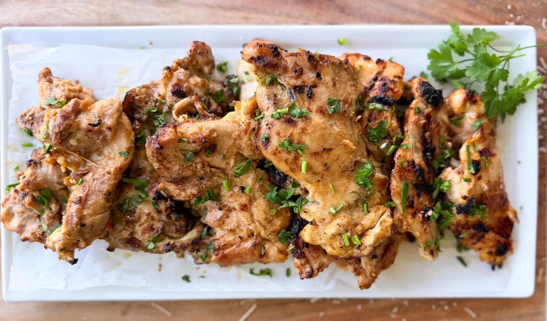 Grilled marinated chicken thighs garnished with chopped herbs, served on a rectangular white plate lined with parchment paper, with a sprig of parsley on the side.