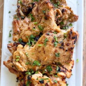 Grilled, seasoned chicken pieces garnished with chopped herbs are arranged on a rectangular white platter with a sprig of cilantro on the side.