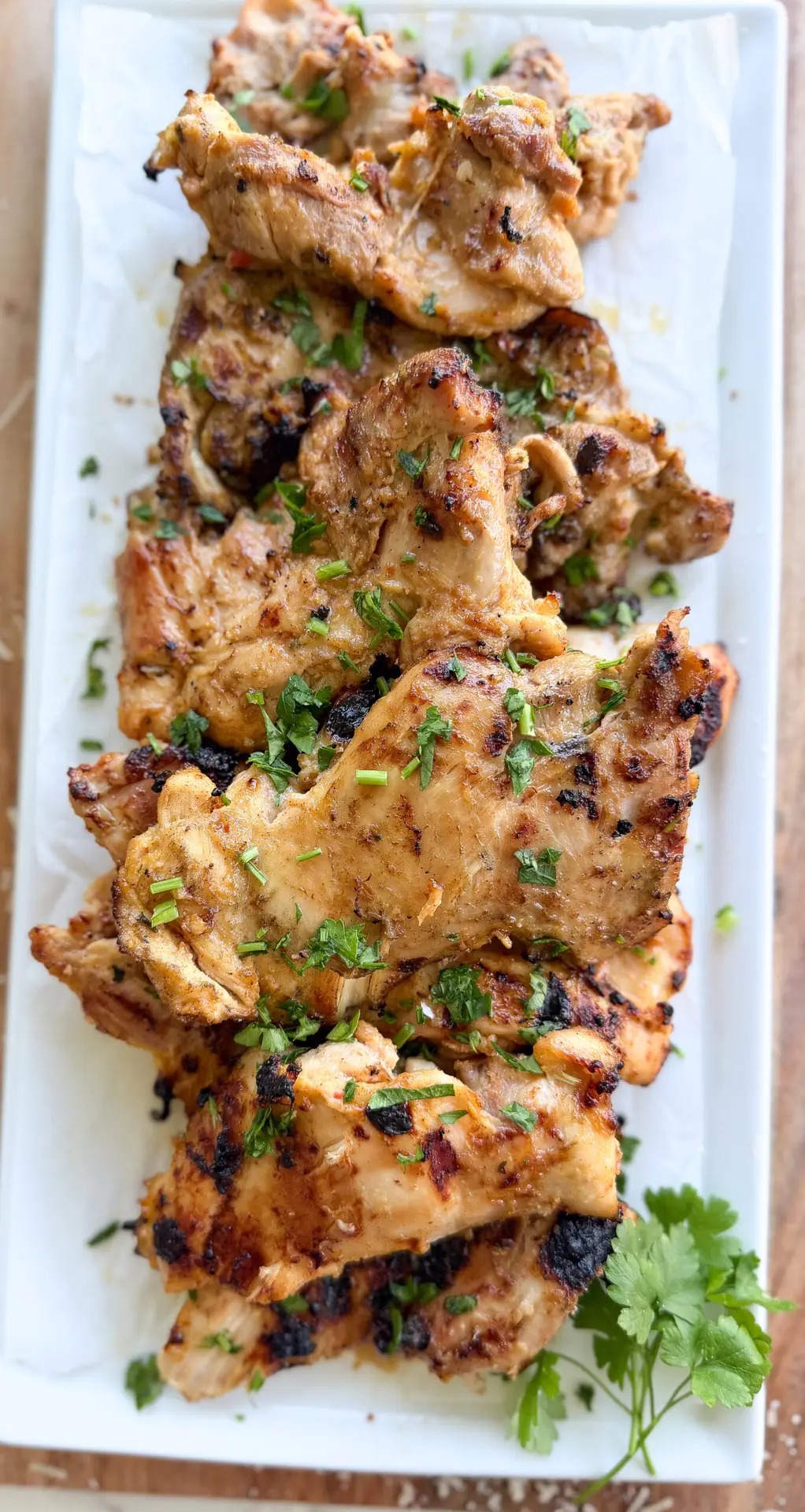 Grilled, seasoned chicken pieces garnished with chopped herbs are arranged on a rectangular white platter with a sprig of cilantro on the side.
