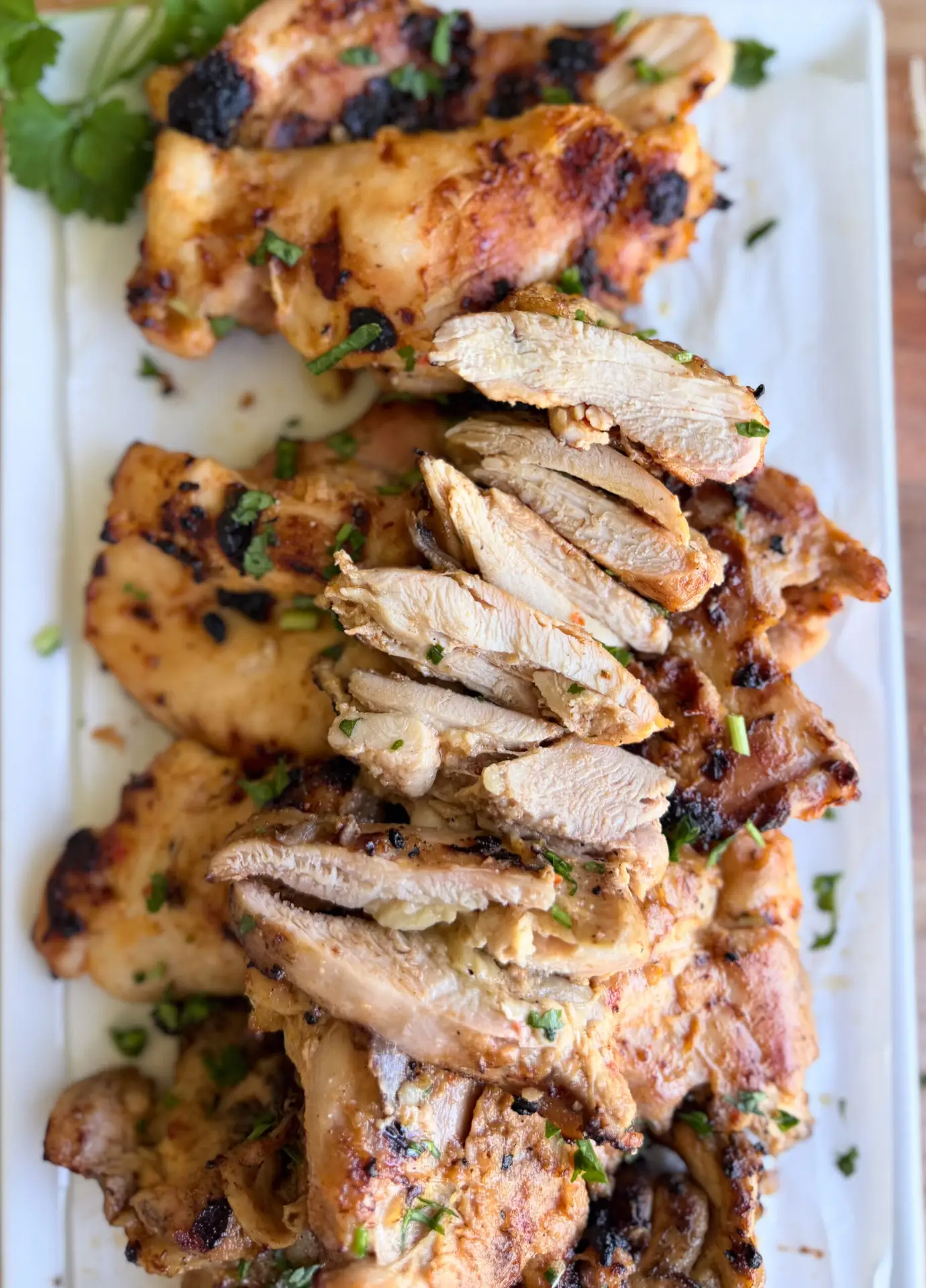 Grilled chicken pieces, some sliced and some whole, garnished with herbs, are arranged on a white rectangular platter.