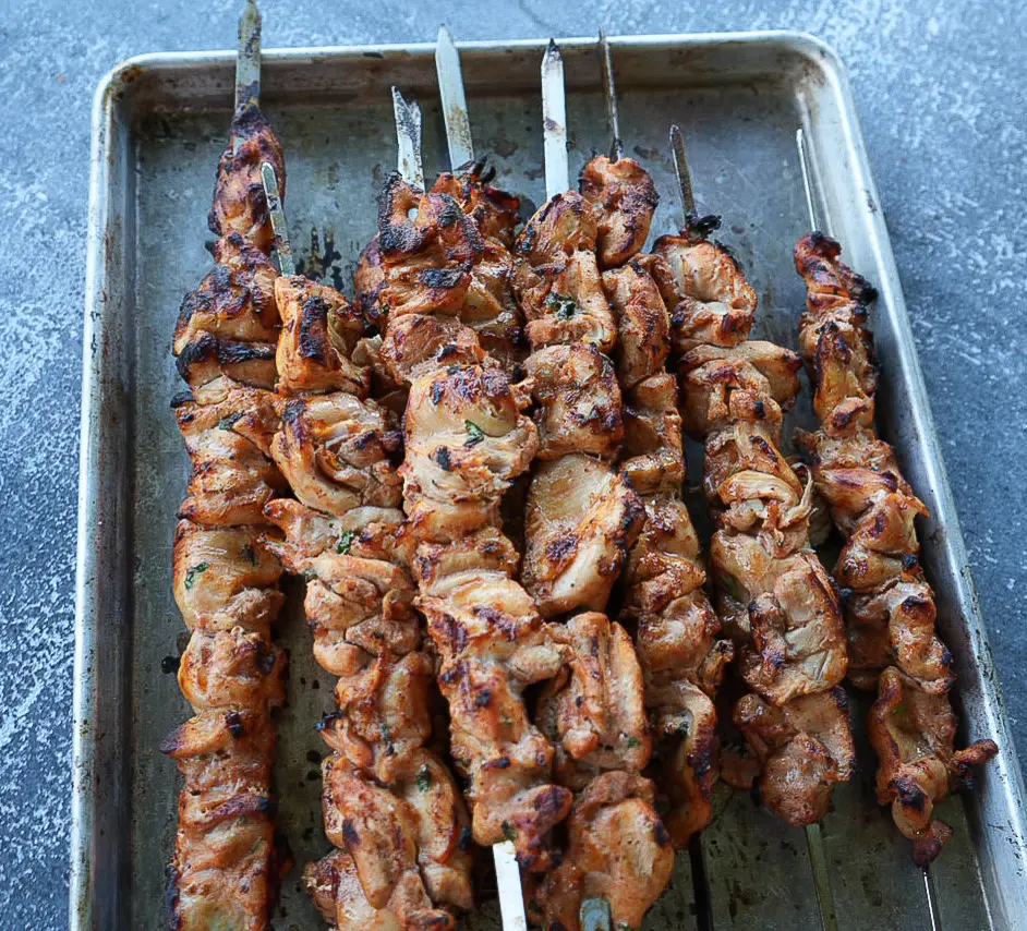 Skewers of grilled marinated meat are arranged on a metal baking tray against a gray surface background.