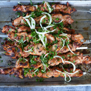 Grilled meat skewers topped with sliced onions and chopped herbs arranged on a metal tray.