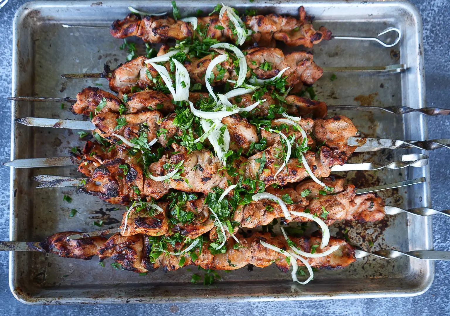 Grilled meat skewers topped with sliced onions and chopped herbs arranged on a metal tray.
