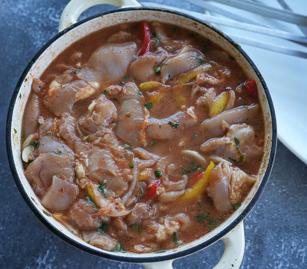 A pot filled with raw, marinated chicken pieces in a reddish sauce with sliced peppers and herbs.