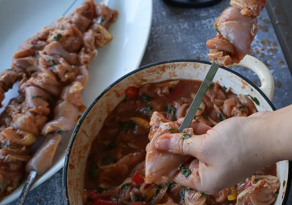 A person skewers marinated pieces of raw chicken over a bowl, preparing kebabs; another skewer with chicken is on a nearby plate.