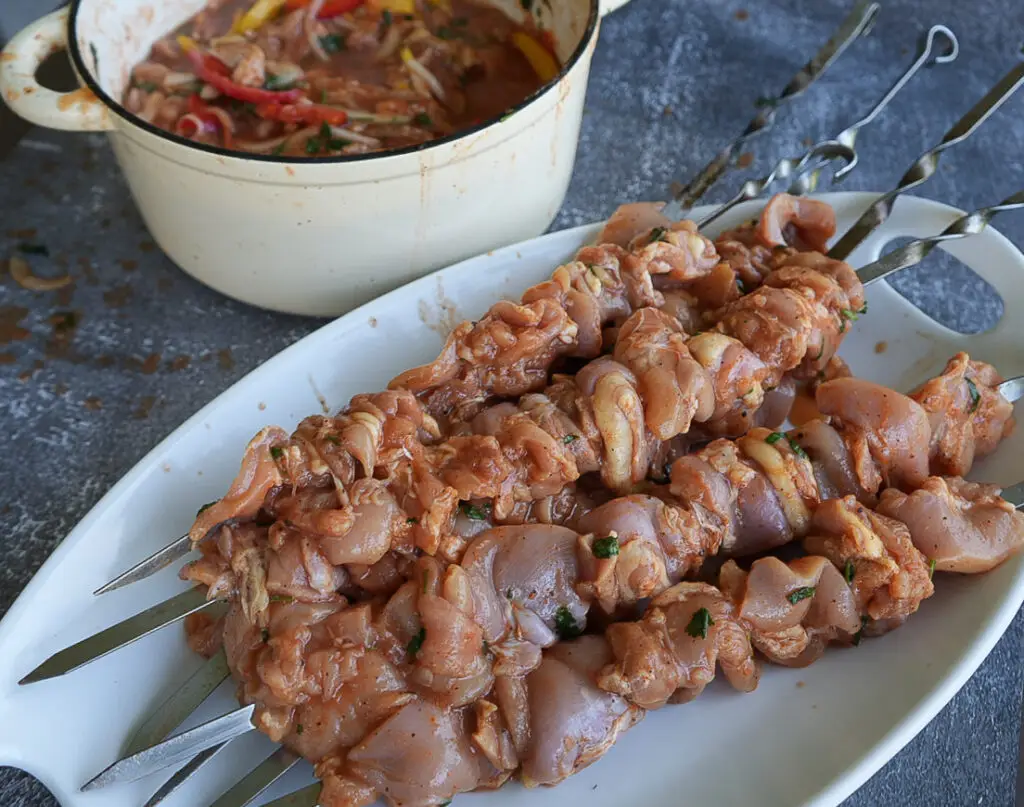 Raw marinated chicken pieces skewered on metal sticks sit on a white plate, with a pot of additional marinade or ingredients in the background.