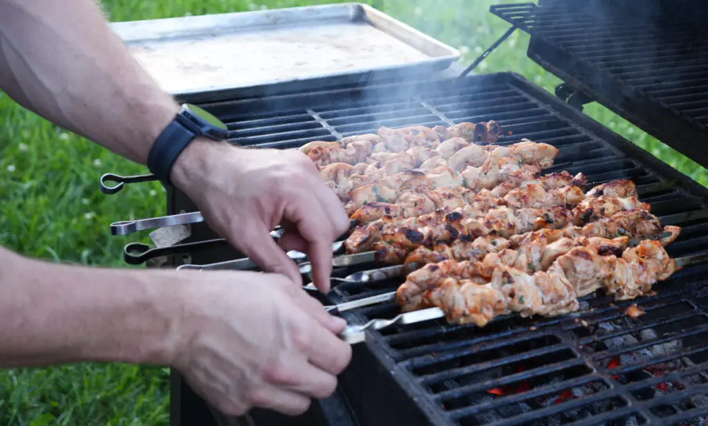A person turns skewers of marinated meat grilling on an outdoor barbecue with smoke rising from the grill.