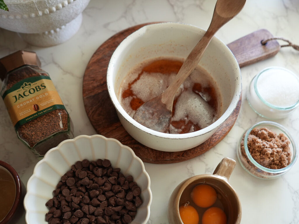 A bowl with melted butter and sugar being mixed, surrounded by chocolate chips, eggs, instant coffee, brown sugar, and white sugar on a marble surface.